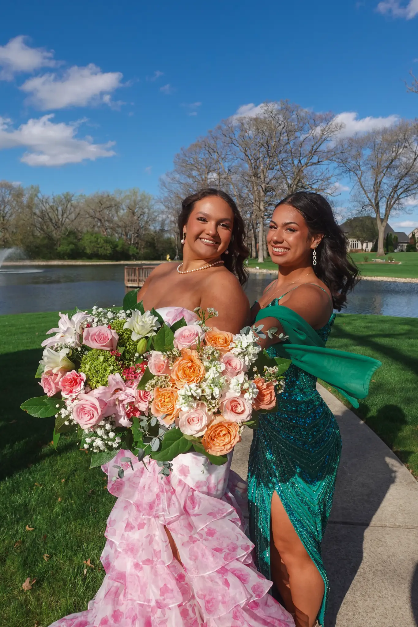 Two prom friends smiling with large bouquets near a lake and blue sky in Northwest Indiana.