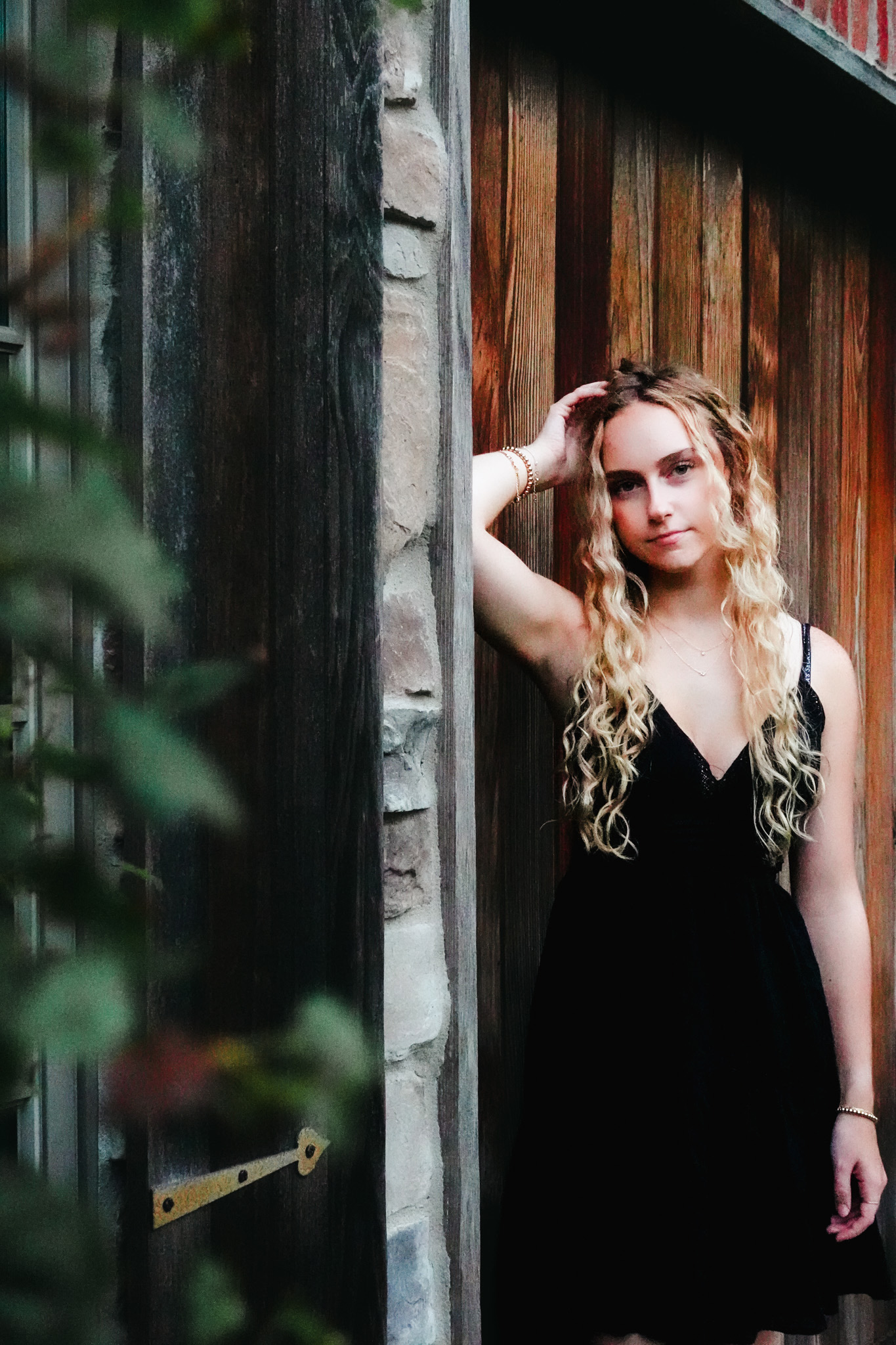 Senior portrait of a blonde client in a black dress standing near a wooden doorway in Northwest Indiana.